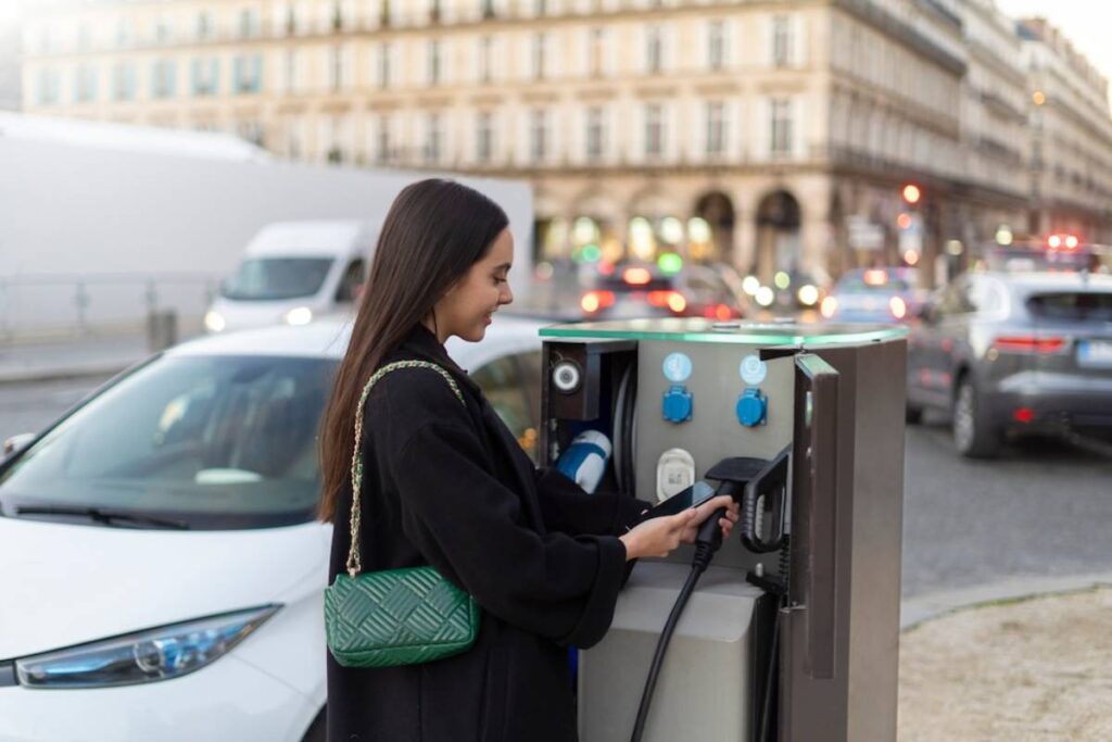 A woman with long hair, wearing a black coat, is charging an electric vehicle at a street charging station.