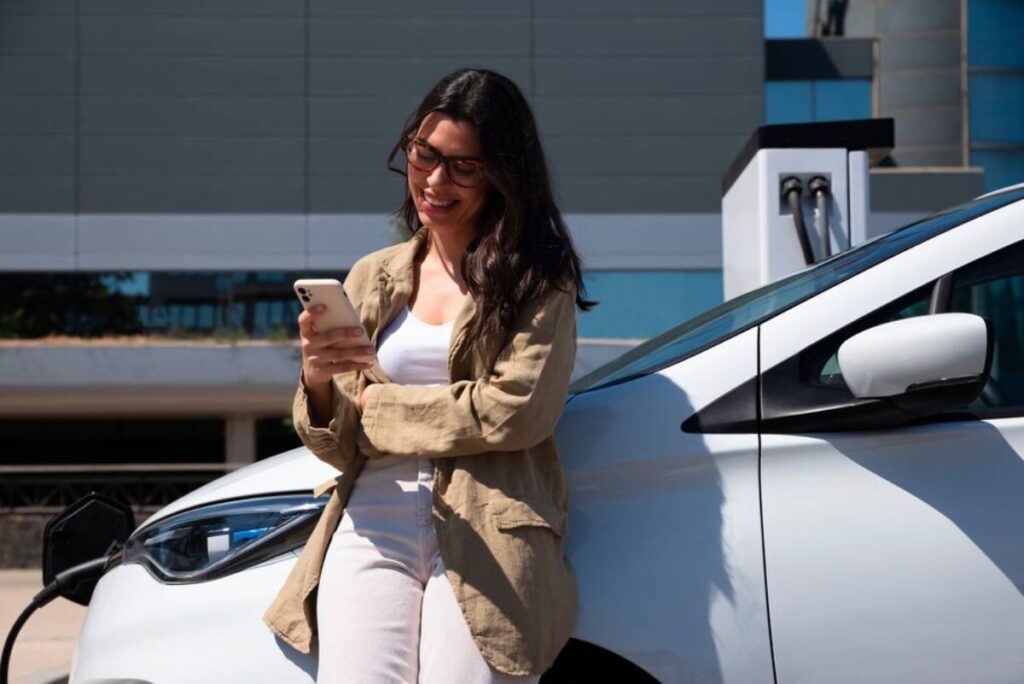 A woman in a beige jacket leans against a white electric car.