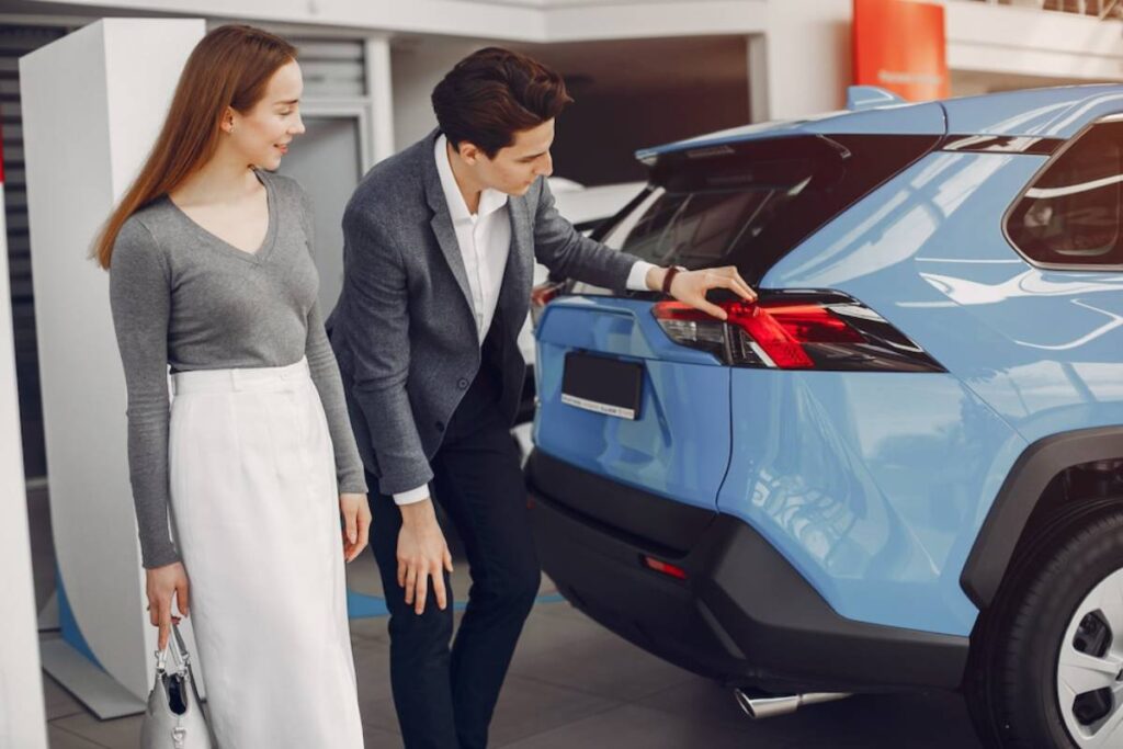 A man in a blazer highlights features of a blue SUV to a woman in a gray sweater.