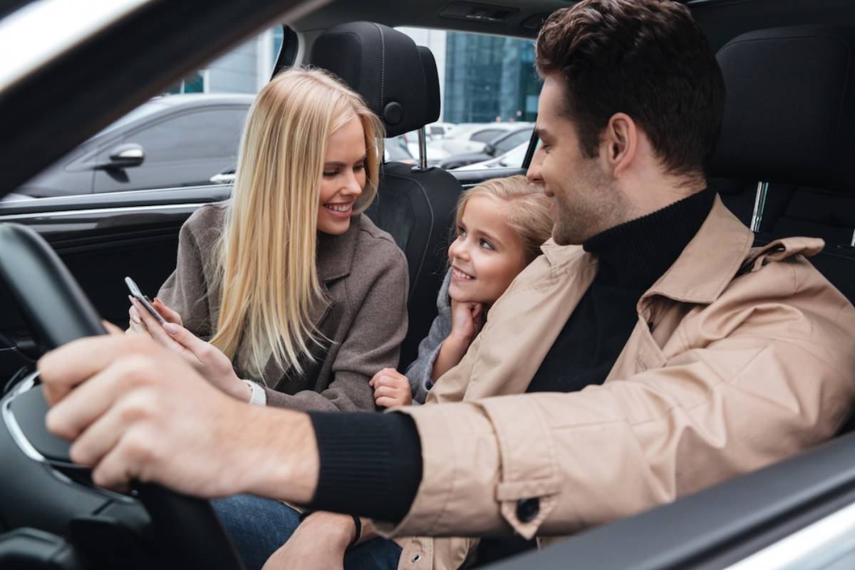   A man and woman sit in a car with their young daughter, all smiling and enjoying a family outing together.
