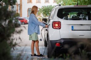 A woman stands beside her electric vehicle, connecting it to a charging station to recharge the car's battery.