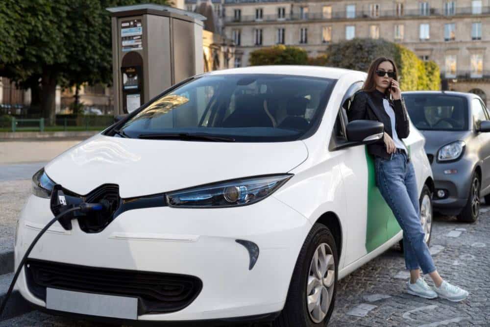 A woman stands beside her electric car, connecting it to a charging station on a city street.
