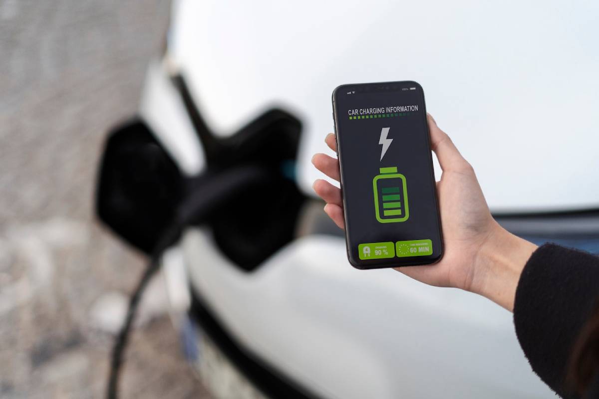  Hand holding a smartphone displaying car charging information, with an electric car plugged in the background.