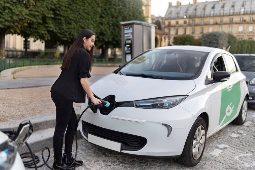  A woman connects a charging cable to an electric car in a parking lot, surrounded by trees and other parked vehicles.
