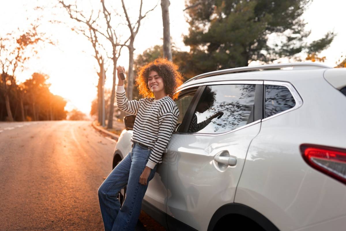 A woman stands beside her car, silhouetted against a vibrant sunset in the background.
