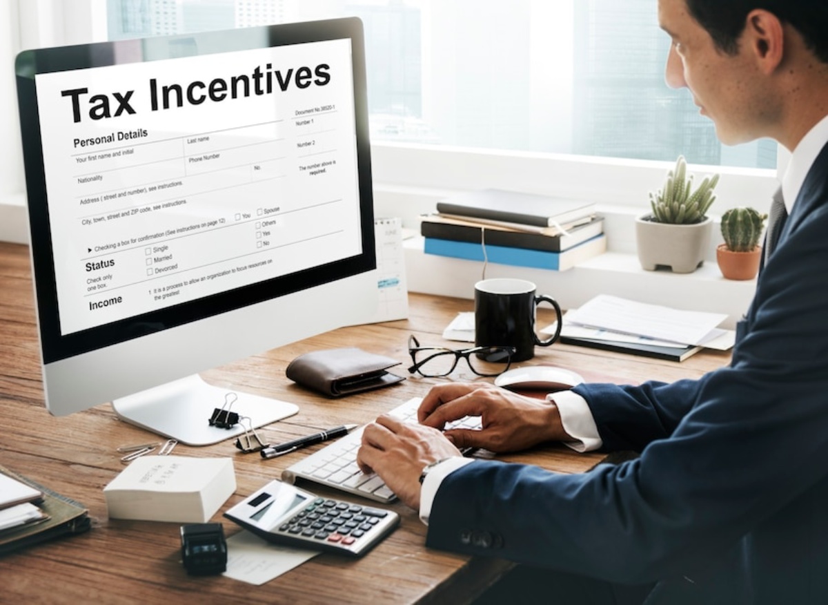 Businessman at desk using computer displaying a tax incentives form, with office supplies and plant in the background.